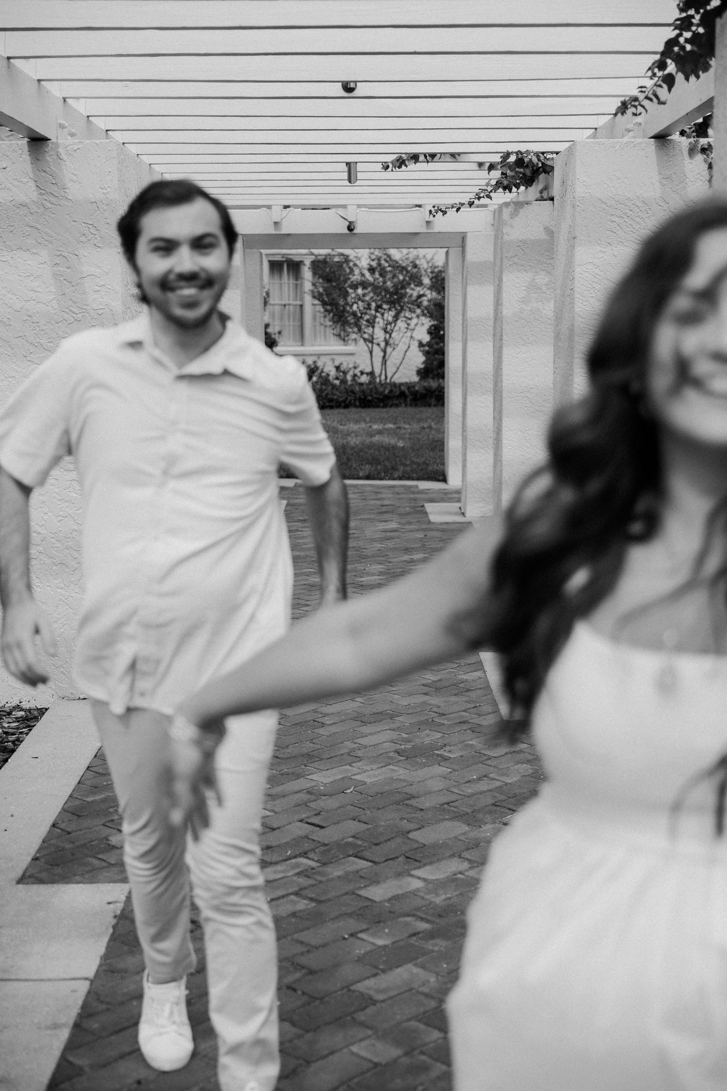 A couple in white attire dances playfully on an outdoor patio with brick pavers during their wedding celebration.