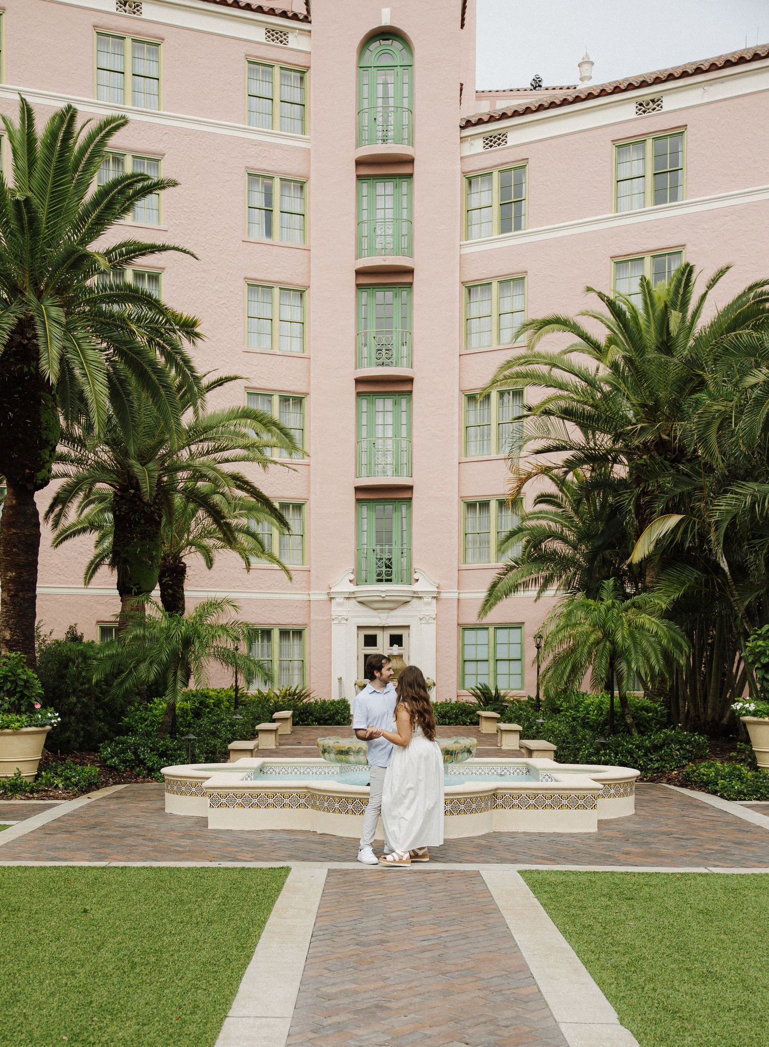 Couple stand together by a courtyard fountain outside a pink Mediterranean-style hotel with palm trees.