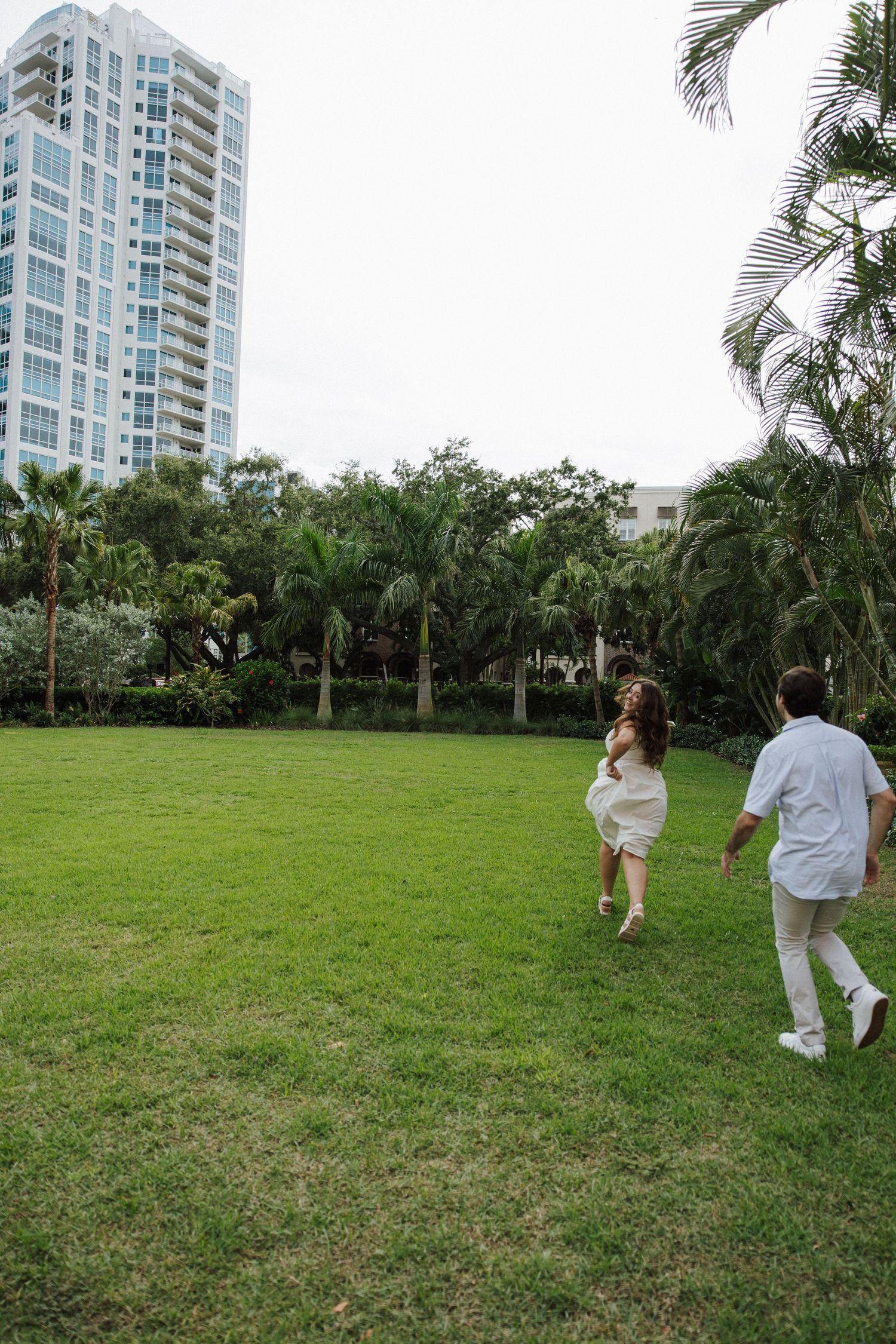 Couple runs playfully across a green lawn near a modern high-rise building surrounded by palm trees.