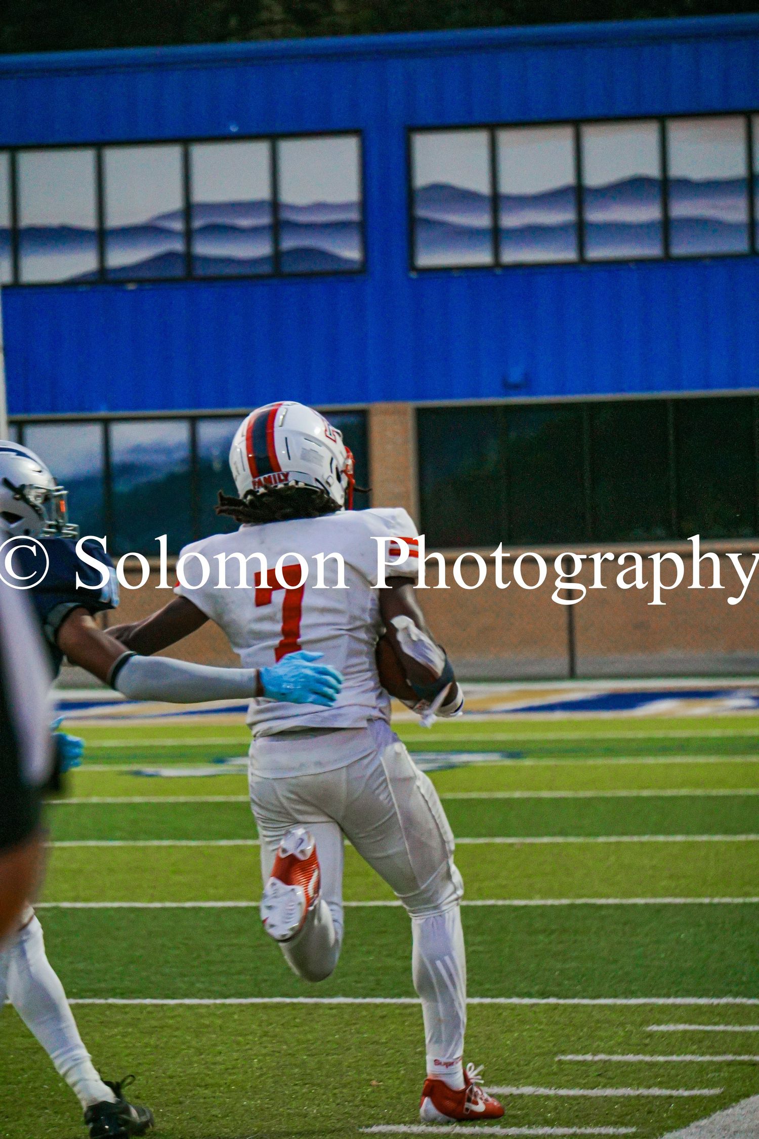 Football player #7 in white uniform runs with the ball during a game captured by Solomon Photography.