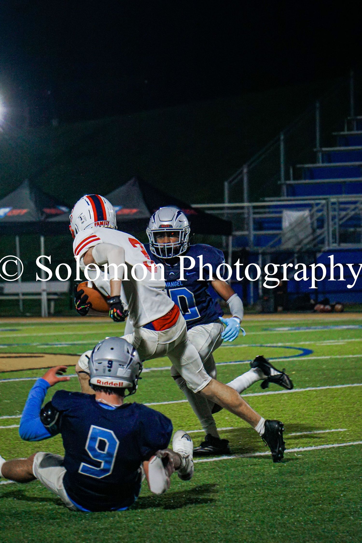 High school football player in orange jersey breaks through two blue-uniformed defenders at night.