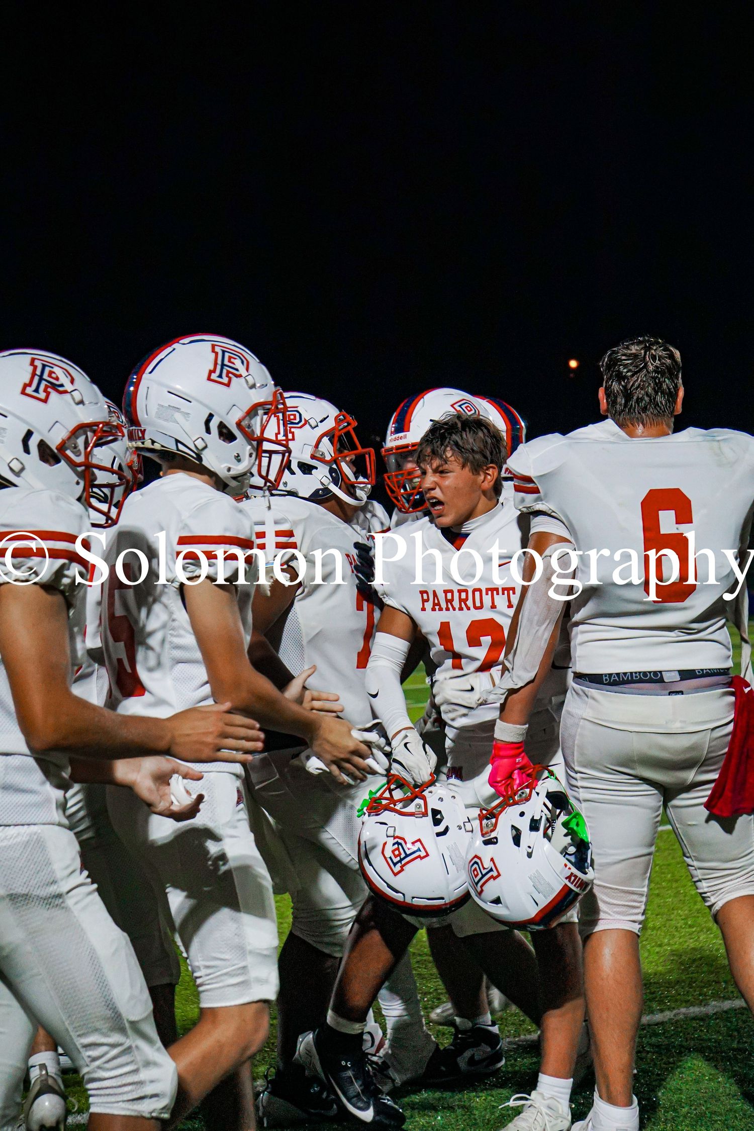 Parrott football team huddles together energetically on the field at night, captured by Solomon Photography.