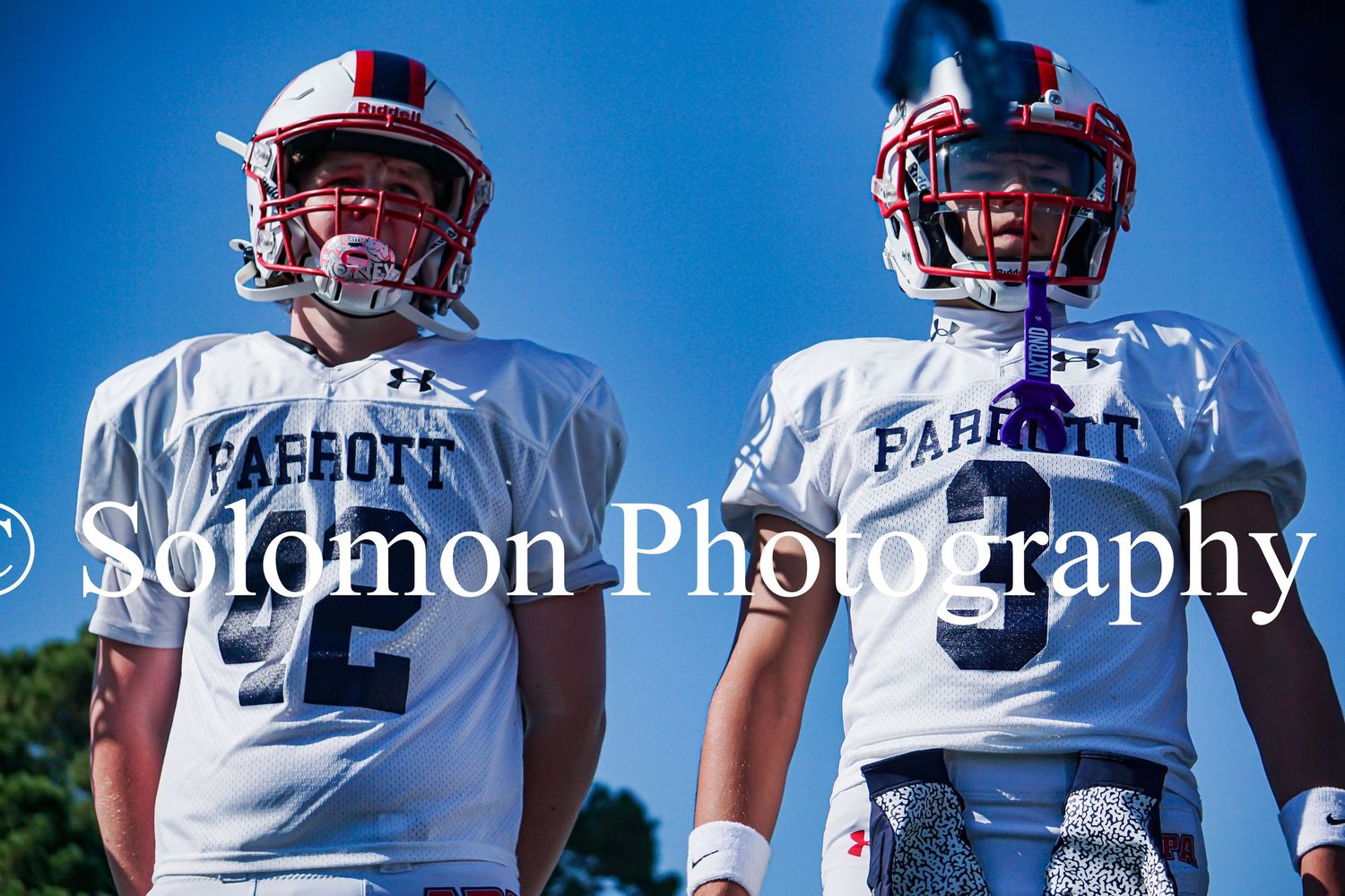 Two Parrott youth football players in white uniforms stand on the field under a blue sky.