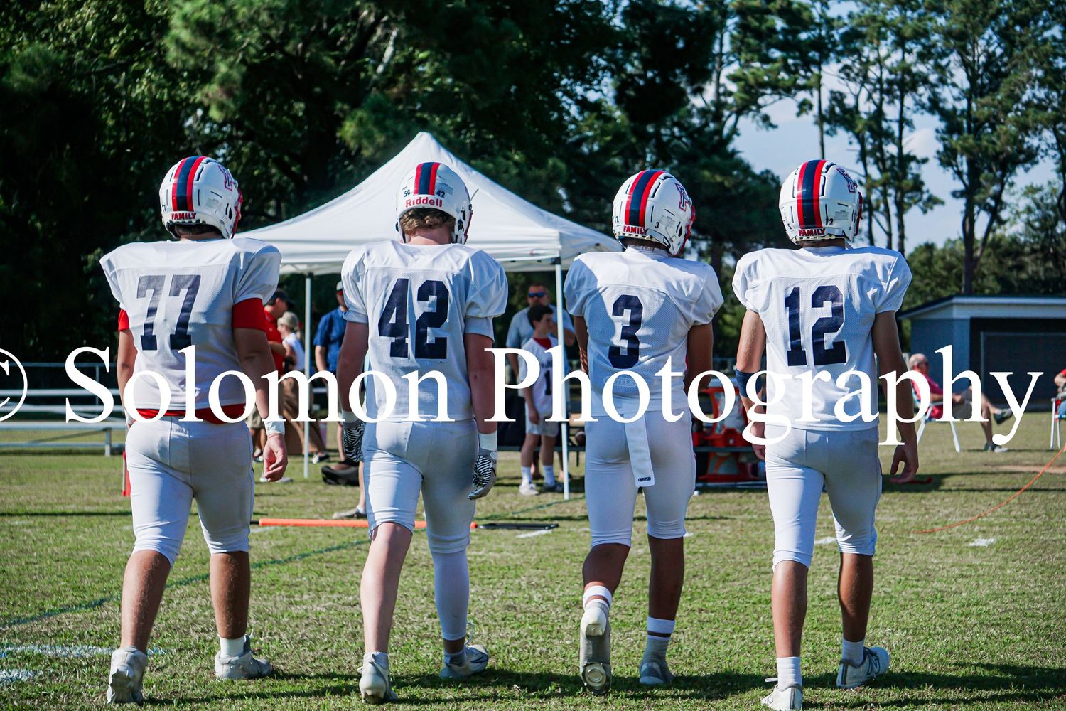 Four Parrott football players in white uniforms with numbers 77, 42, 3, and 12 stand on the sideline.