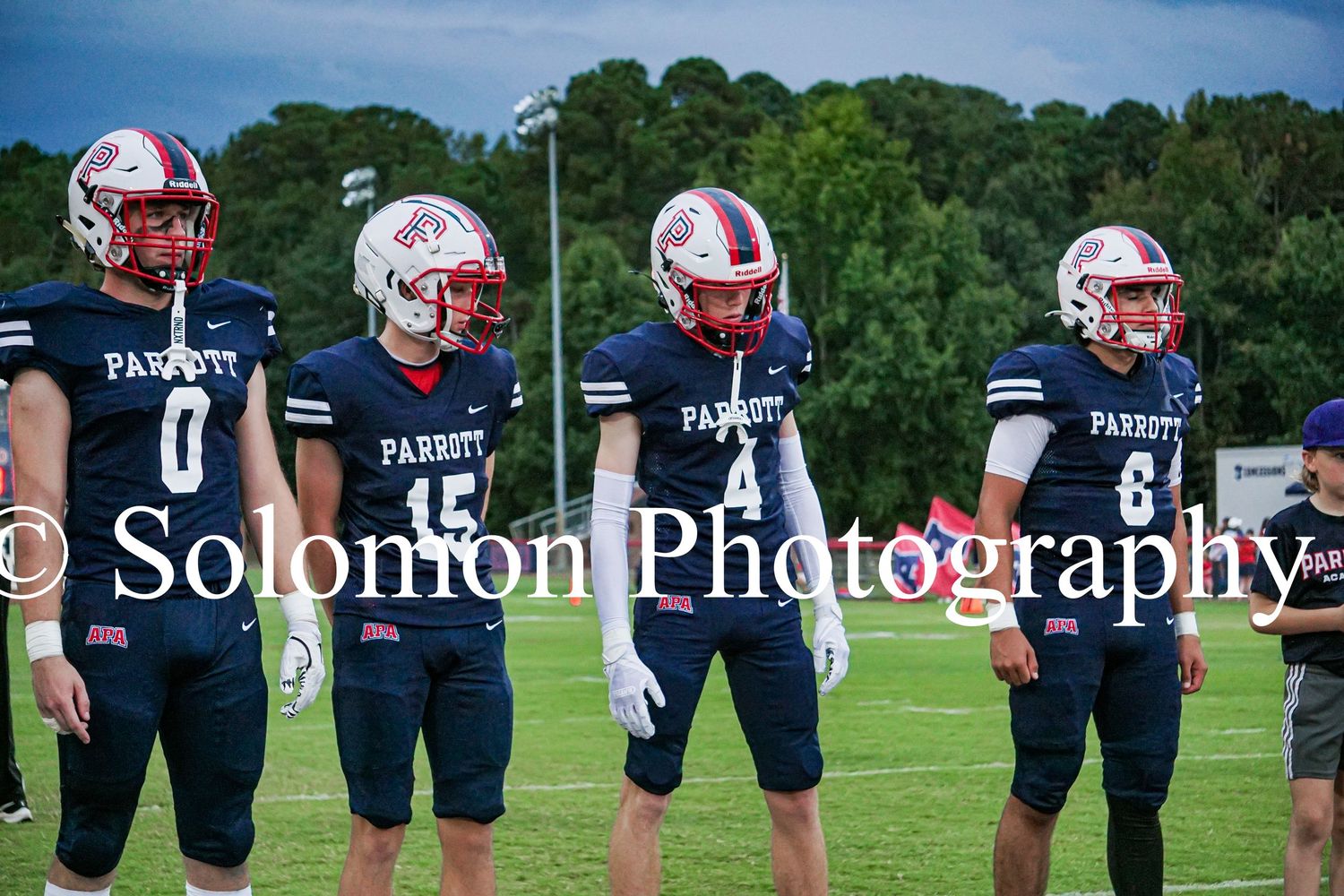 Four Parrott Academy football players in navy uniforms and red helmets stand on the sideline before a game.