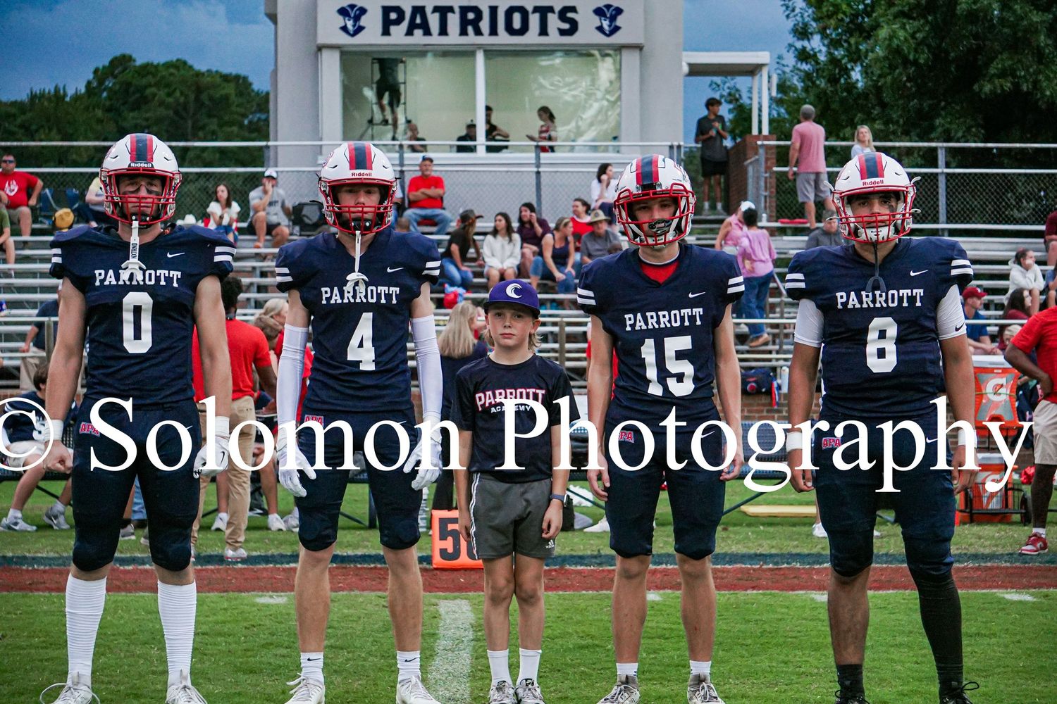 Parrott Patriots football players in navy jerseys pose with a young child on the field before a game.