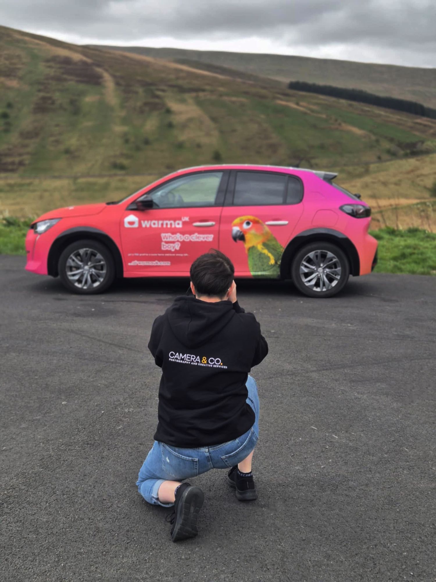 Pink branded vehicle photographed against rural hillside with photographer kneeling in foreground.