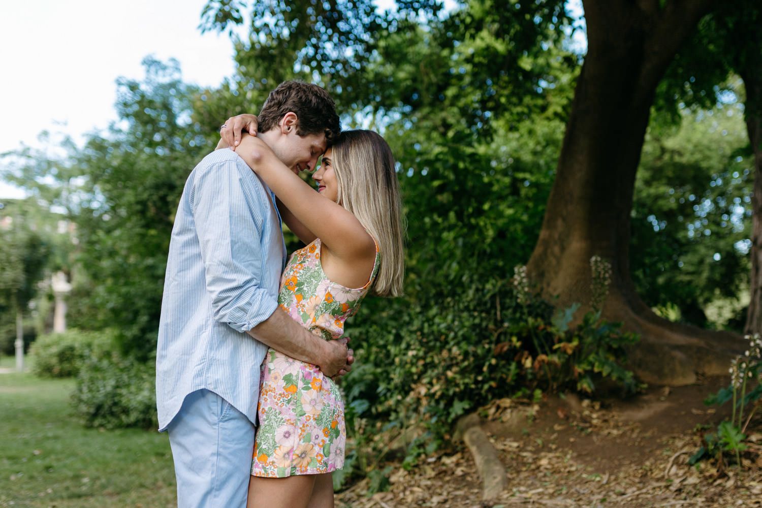 Romantic couple embraces under lush green trees in an outdoor garden setting.