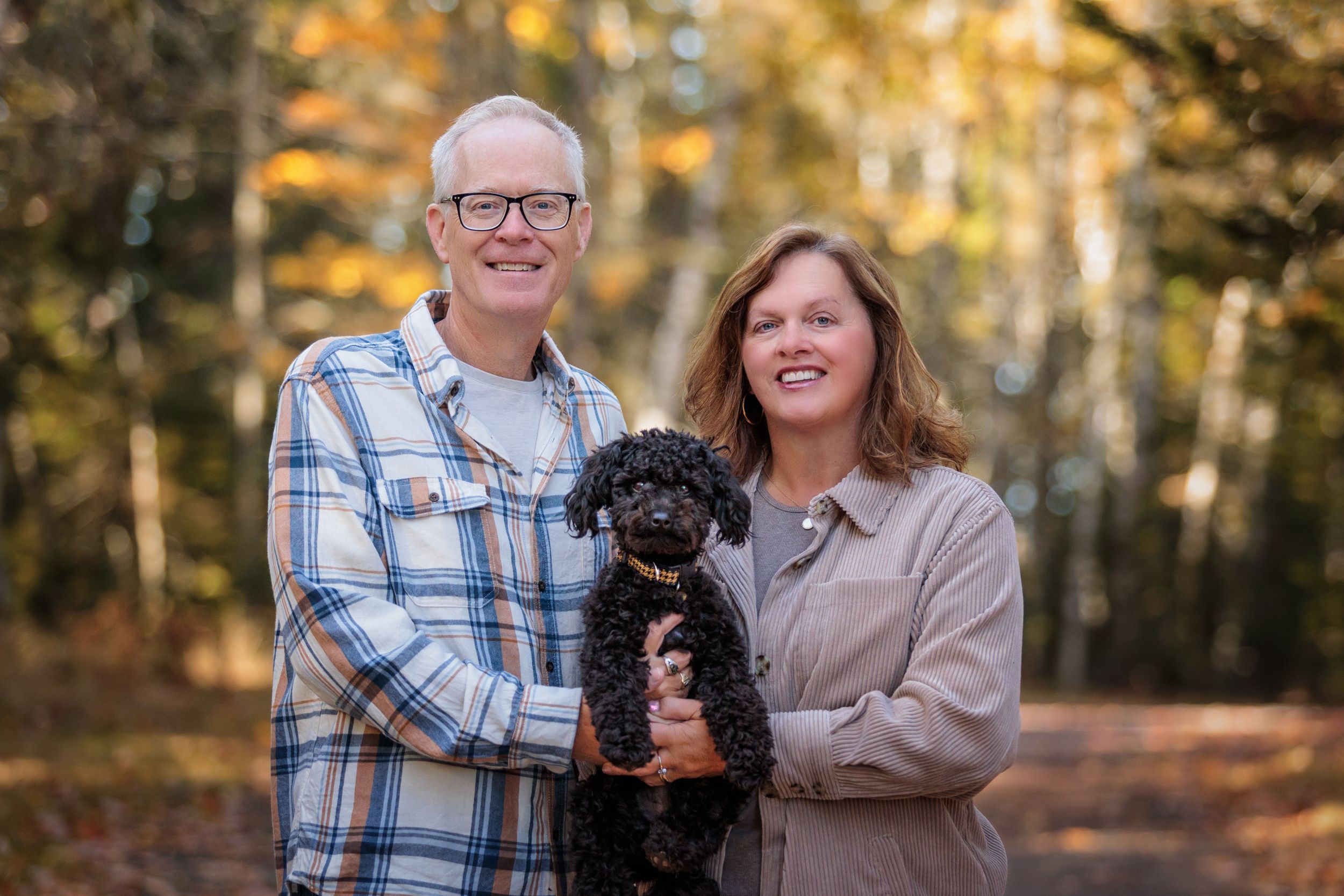 A couple in plaid and khaki hold their small black dog while posing outdoors during golden autumn light.