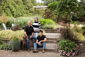 Three people pose with a brown dog on stone garden steps surrounded by flowering plants, tall grasses, and a wooden pergola.