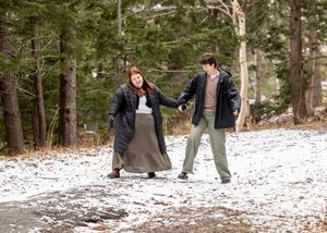 A couple walks hand in hand on a snowy forest path wearing winter coats and warm clothing.