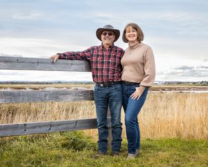 A couple in casual attire stands close together in golden grassy field during sunset with ocean views.