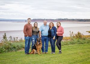 Family of five posing with a German Shepherd on a grassy shoreline overlooking calm water and a distant tree-lined coast under a cloudy sky.