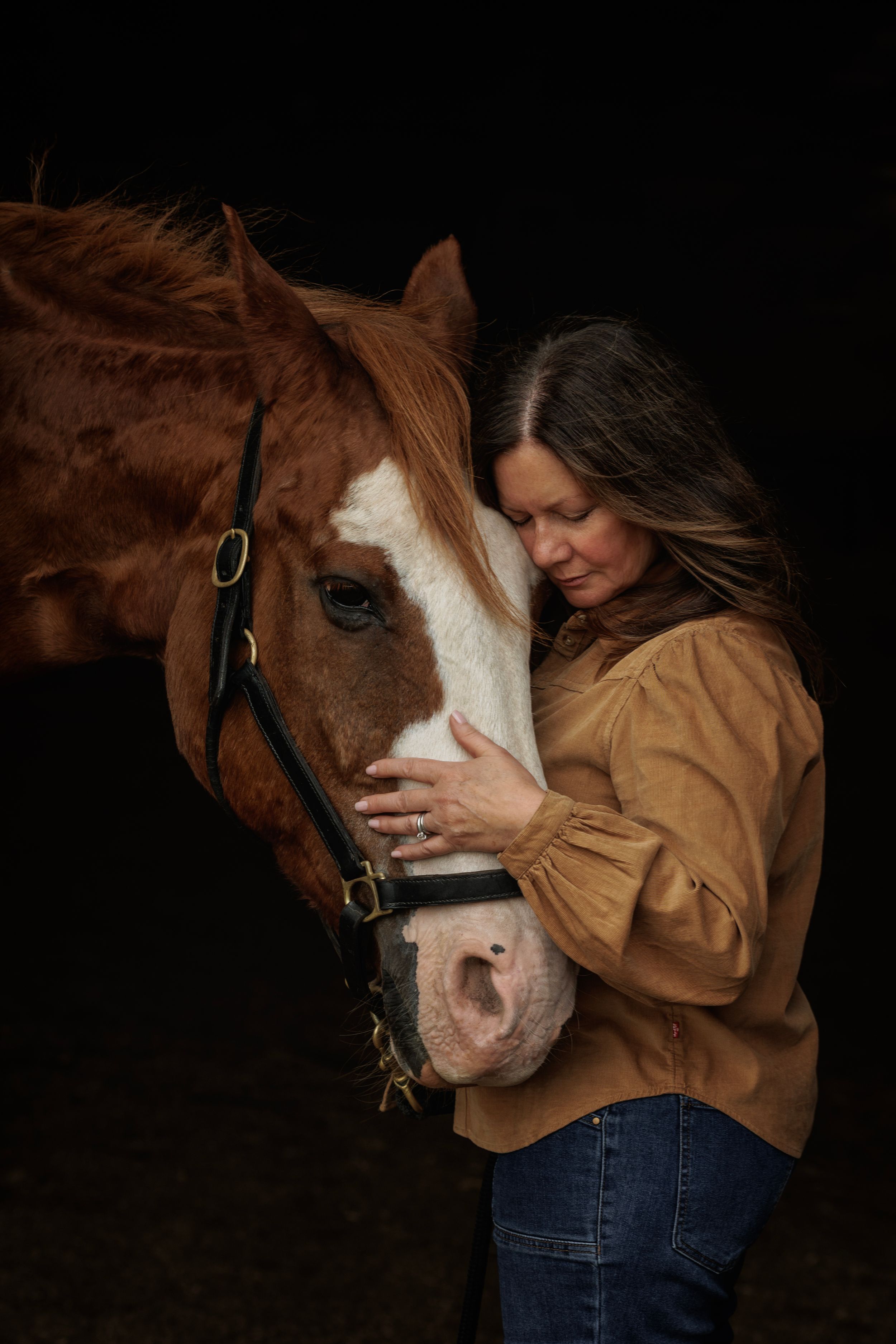 A woman in a tan blouse and jeans gently embraces a chestnut horse with a white blaze against a dark background.