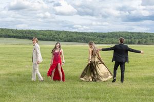 Four young people in formal prom attire, including a red dress and a gold gown, walk across a wide green grass field under a cloudy sky.