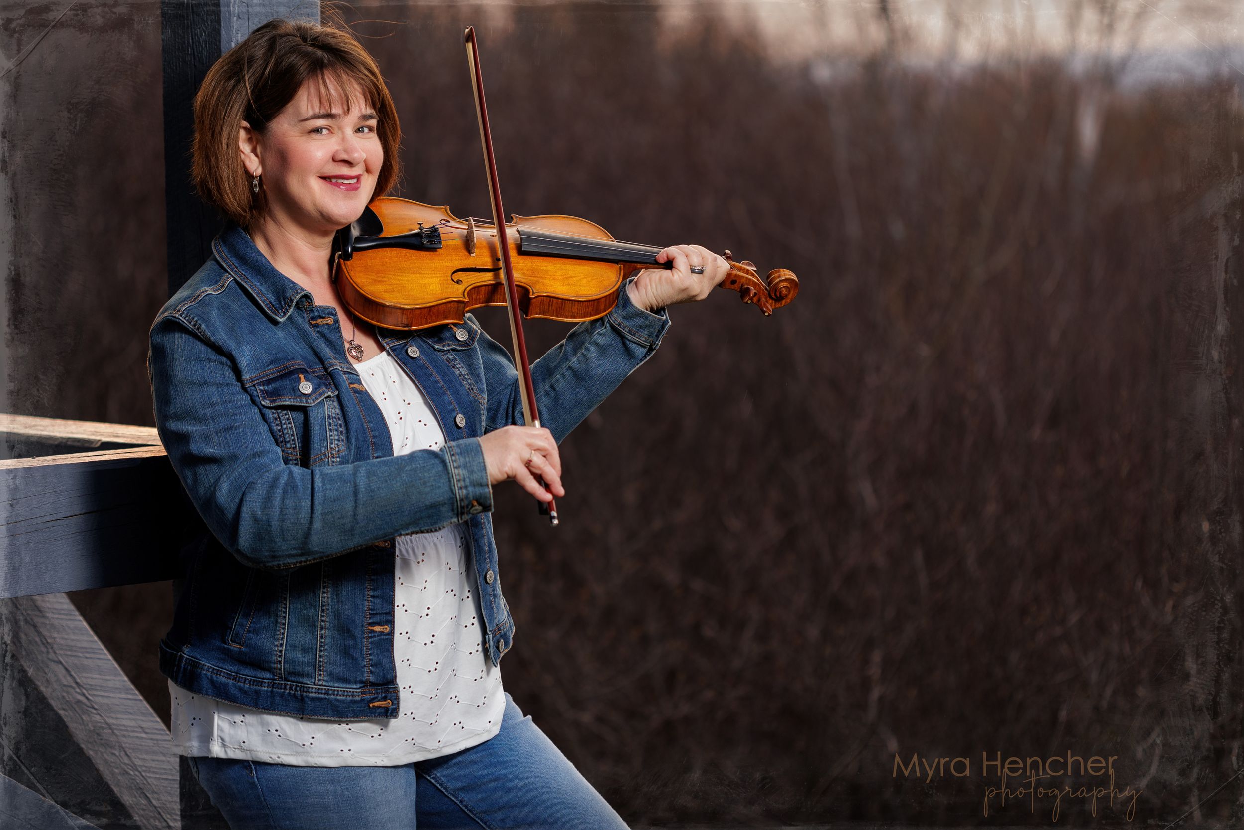 Musician in denim jacket plays violin with enthusiasm against dark background.