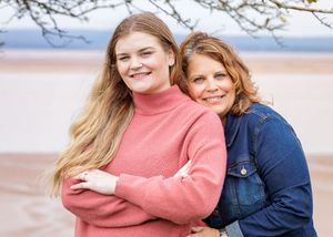 Two smiling women pose together outdoors near a beach, one in a pink sweater and one in a denim jacket.