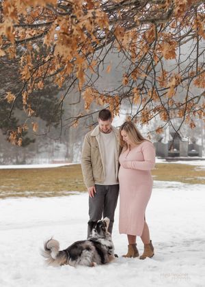 A couple stands in a snowy park under orange autumn leaves with their husky dog laying in the snow.