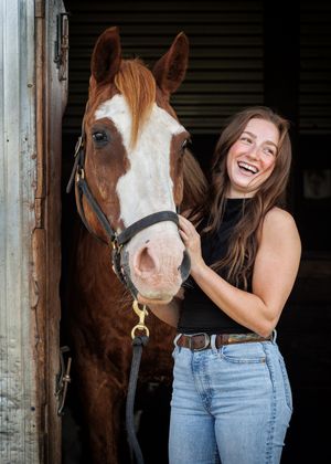 Smiling young woman with long brown hair laughs while standing beside a chestnut and white horse in a dark barn doorway.