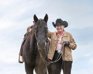A person in western attire stands with a dark horse against a light sky while wearing a tan jacket and cowboy hat.