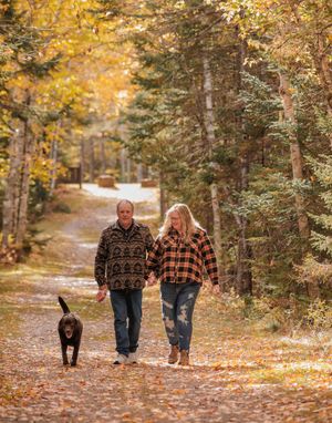 Couple holding hands walks a black dog along a leaf-covered path through colorful autumn trees.