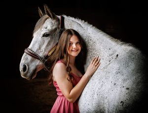 A person in a pink dress stands next to a beautiful white horse in a dark indoor setting.