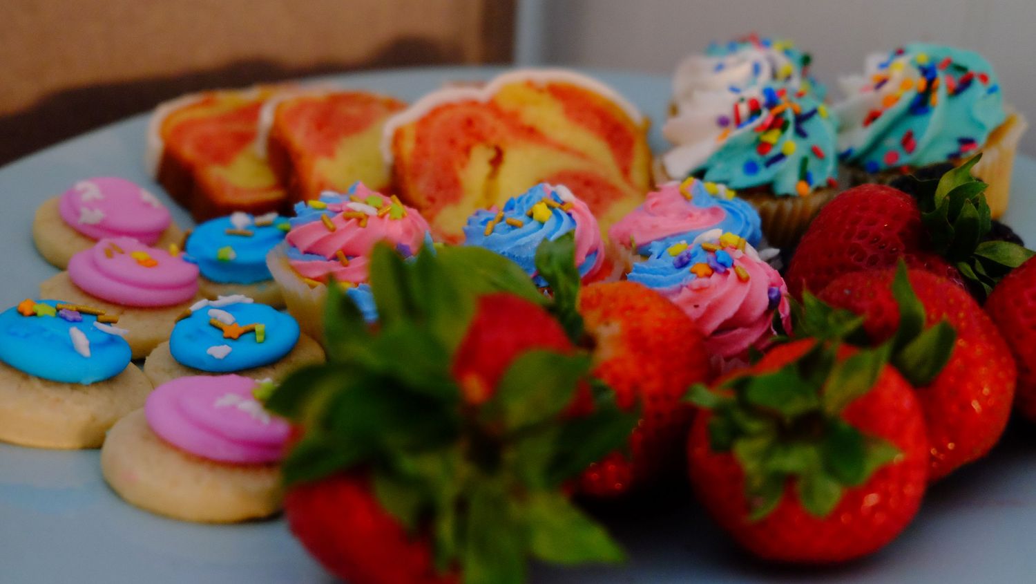 Colorful frosted cookies with red strawberry halves and mint leaves arranged in neat rows on a wooden surface.