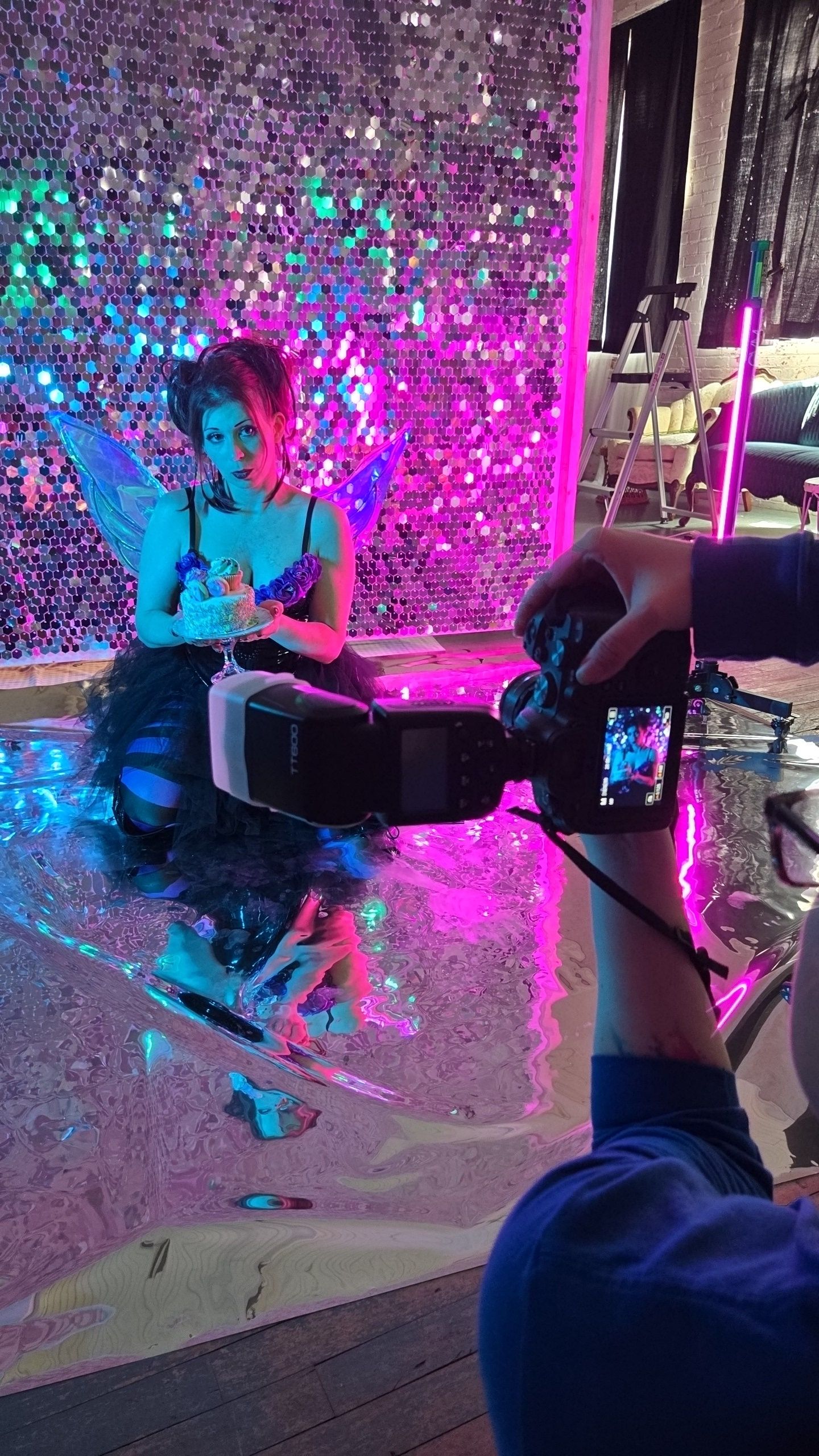 Behind-the-scenes photoshoot with colorful pink and blue lighting against a sparkly sequined backdrop.