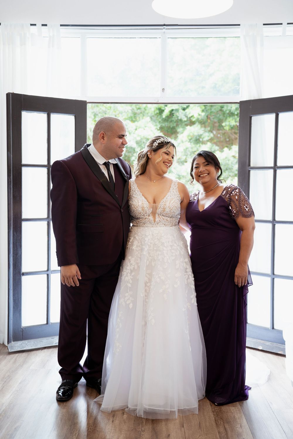 A wedding party in formal attire stands together in a sun-lit venue with a bride in a white gown and guests in dark dresses.