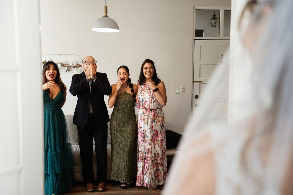 Wedding guests react with joy and emotion while viewing someone in an elegant white room interior.