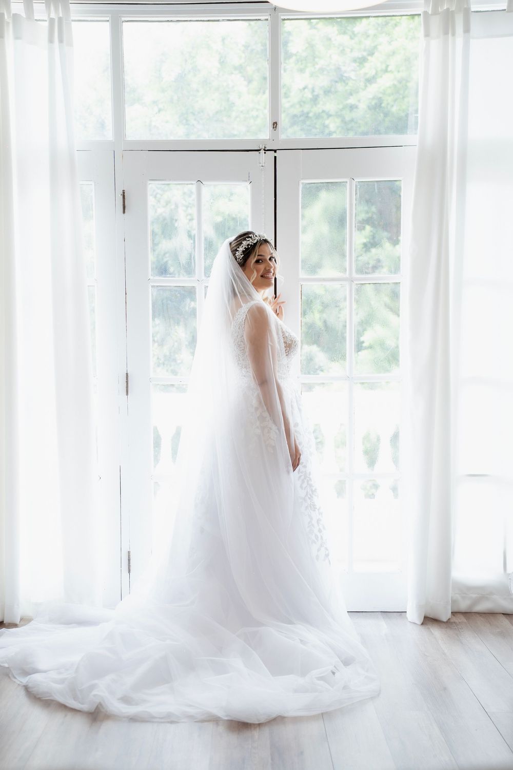 A bride in a flowing white wedding gown and veil stands elegantly by sunlit windows with sheer white curtains.