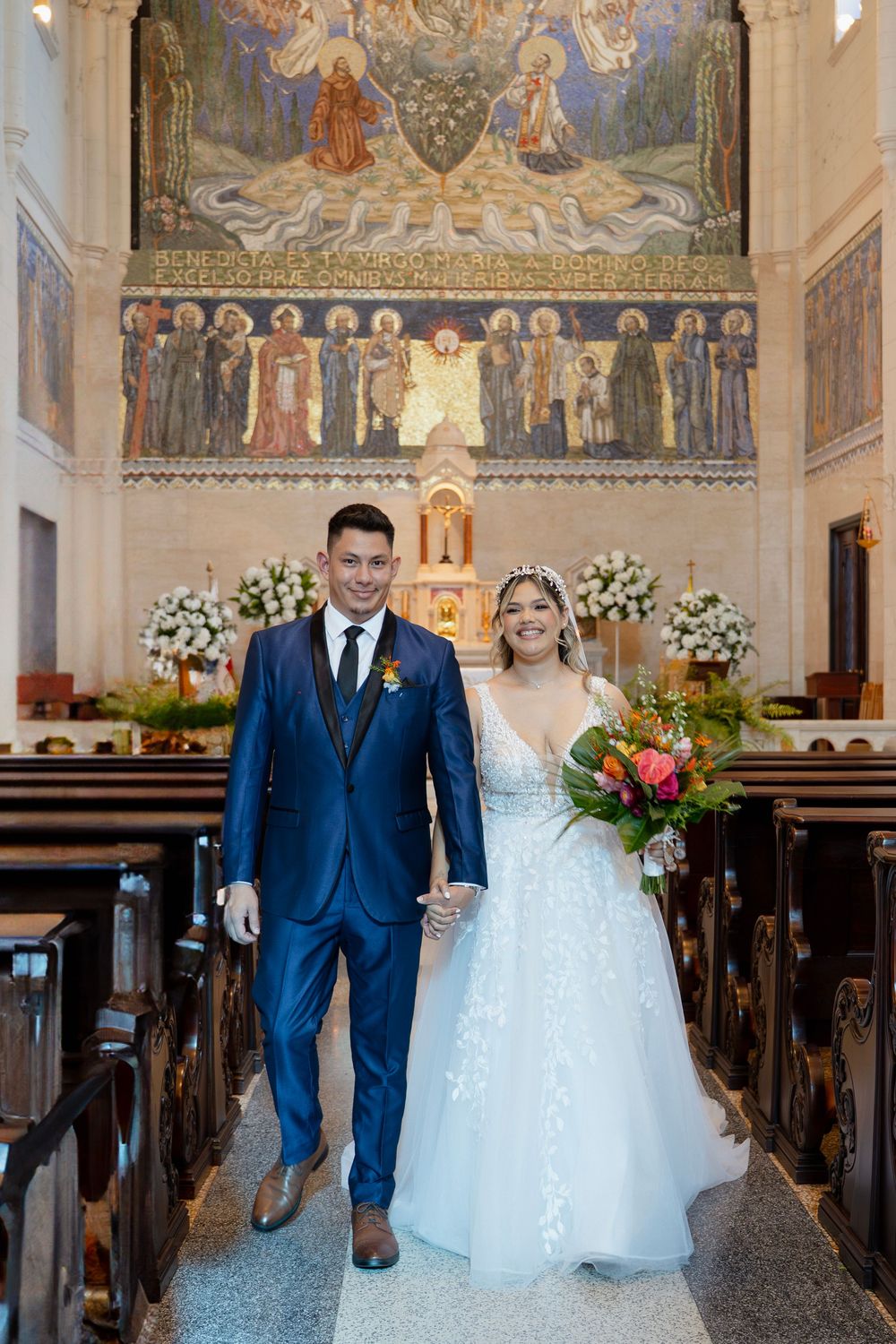 Married couple poses in church aisle with traditional Catholic artwork and altar decorations visible in background.