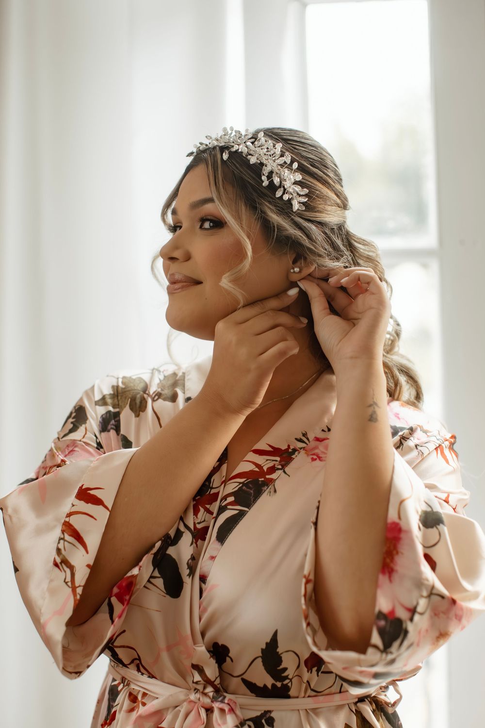 Bride getting ready in a floral silk robe while wearing a delicate crystal hair crown against a bright window.