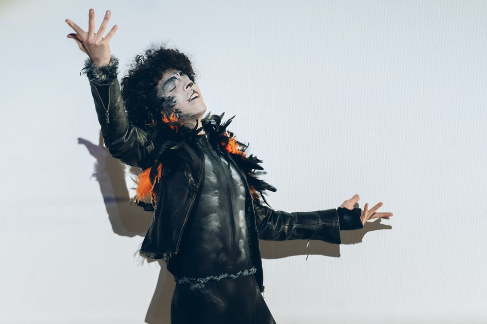A performer in a black leather jacket and feather boa strikes a dramatic pose against a white background.