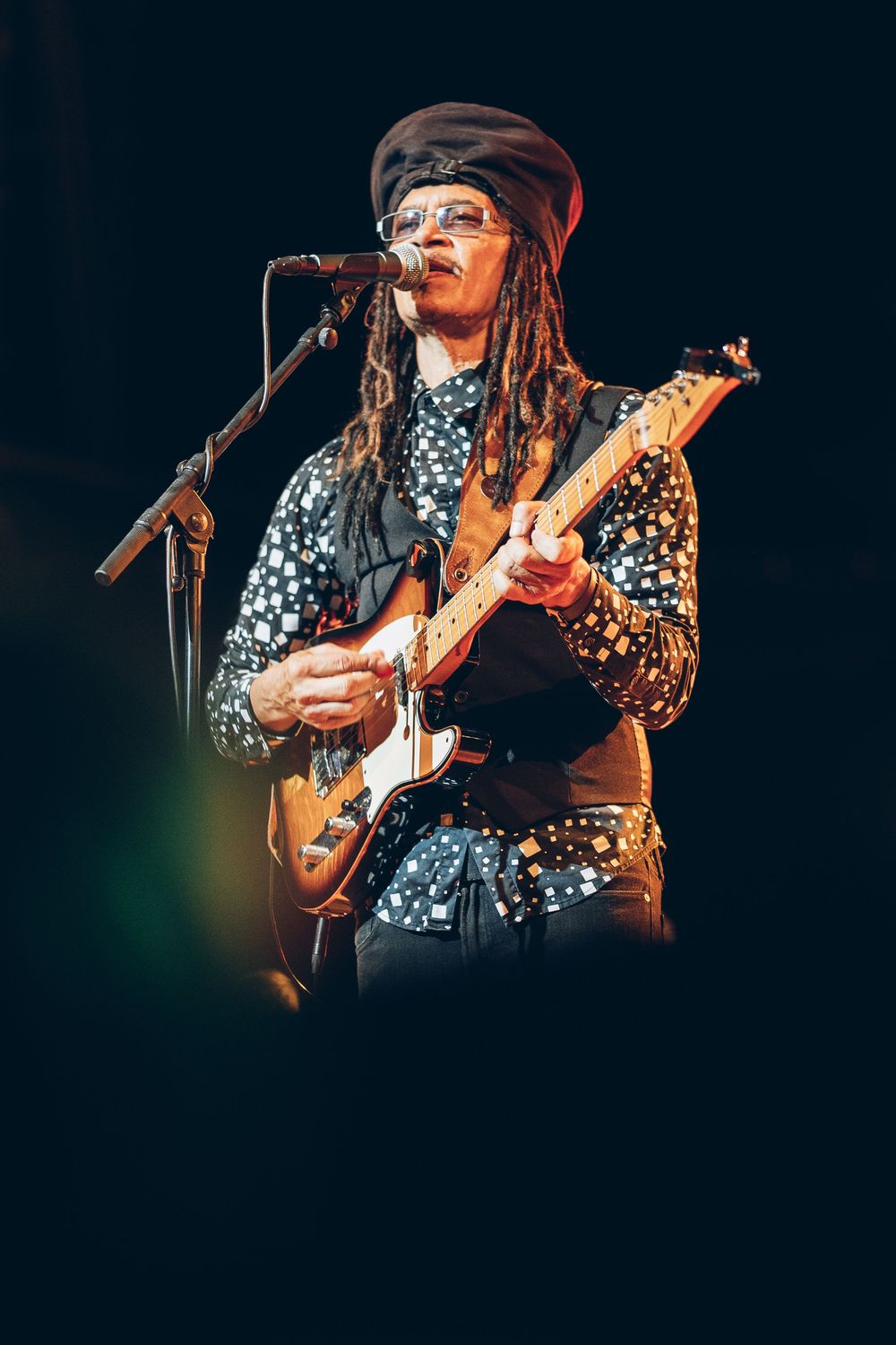 Musician performs on stage wearing a floral print shirt and hat while playing guitar under dramatic concert lighting.