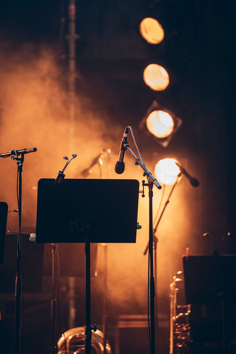 Silhouetted music stand and microphone stands on stage with warm orange stage lights glowing in the background.