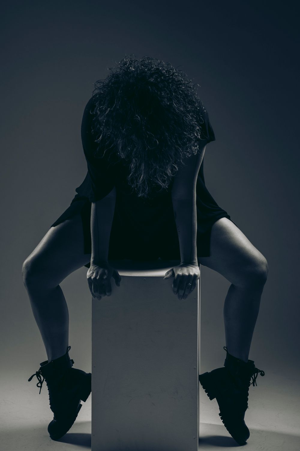 Dark silhouette figure wearing boots sits on white cube in moody artistic portrait against gray background.
