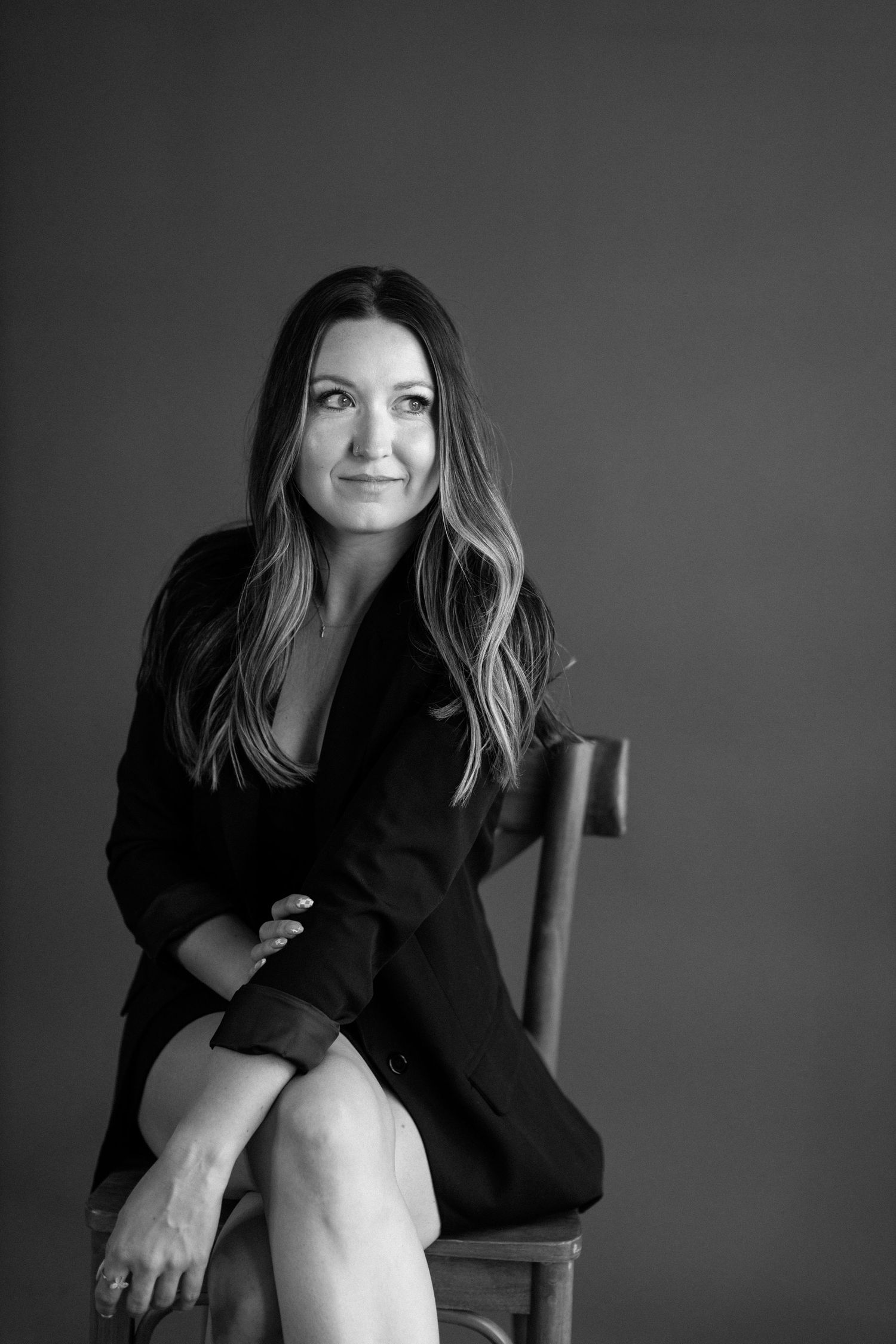 Professional black and white portrait photograph of someone in a black blazer sitting on a wooden chair against a dark background.