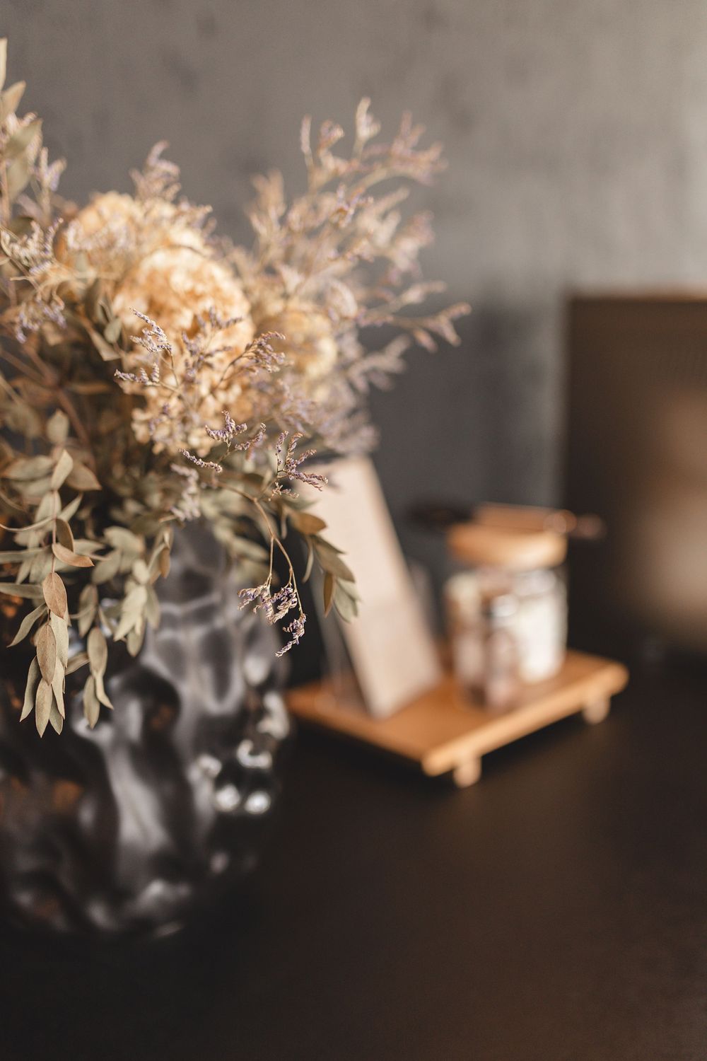 Dried floral arrangement in a dark glass vase on a black table with a wooden menu holder in the background.
