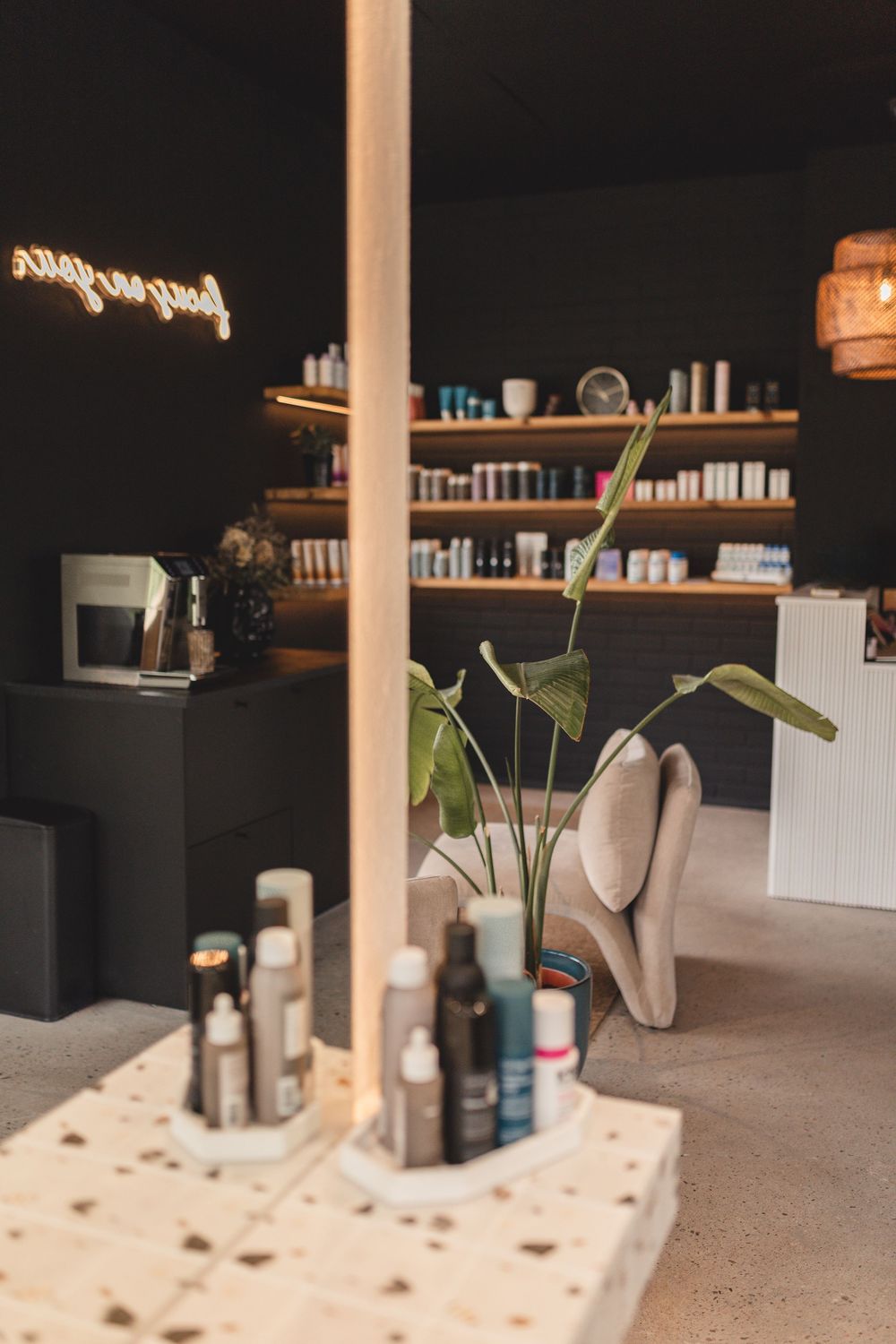 Modern hair salon interior with dark walls, wooden shelves displaying hair care products, and a tropical plant centerpiece.