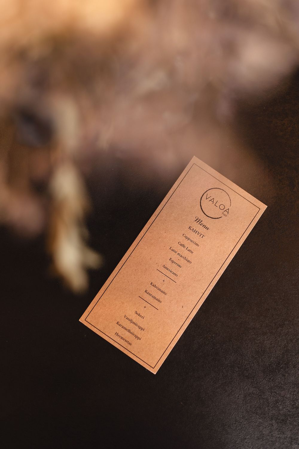 Kraft paper menu card for Valoa restaurant lying on a dark surface with blurred floral background.