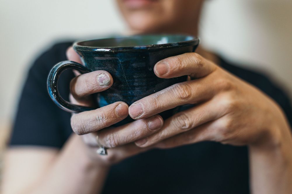Close-up of clay-smudged hands holding a dark teal handmade ceramic mug with a rich glazed finish.