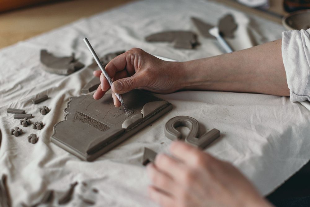 Hands carving clay letters and shapes on a fabric-covered work surface in a pottery studio.
