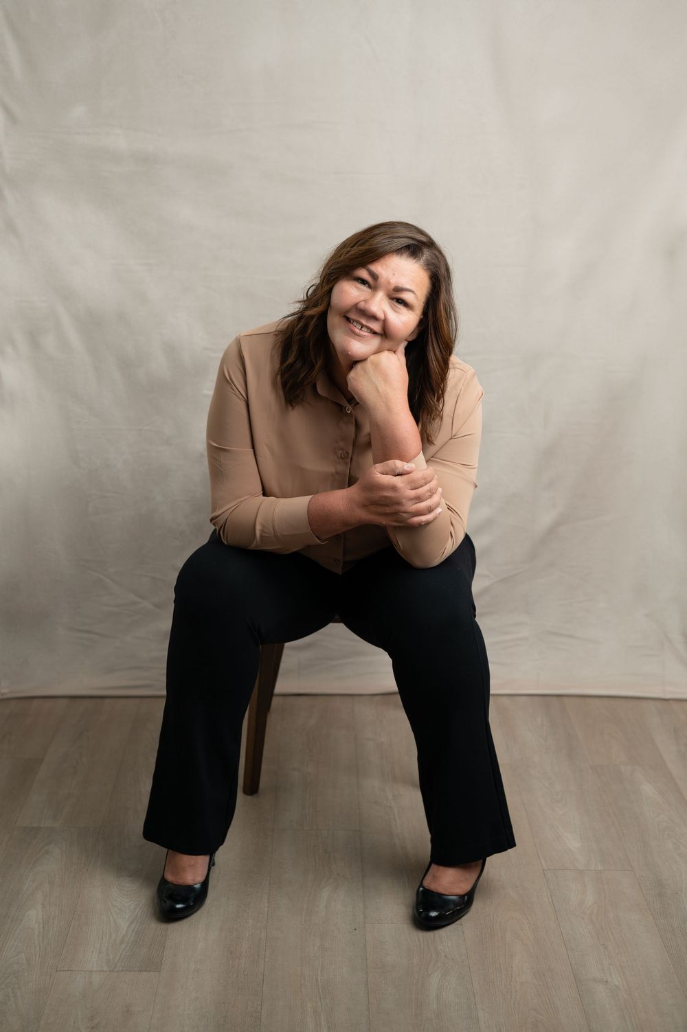 A person wearing a beige sweater and dark pants sits casually on a chair against a light backdrop with a warm smile.