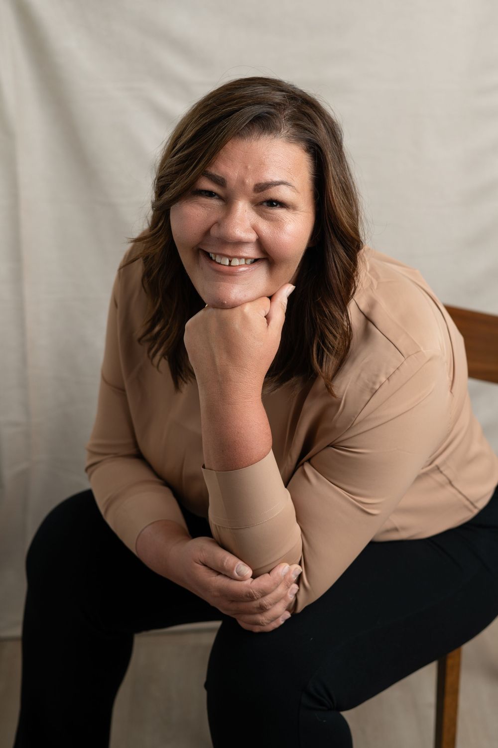 Professional indoor portrait of someone in a beige top and black pants sitting casually on a chair against light backdrop.