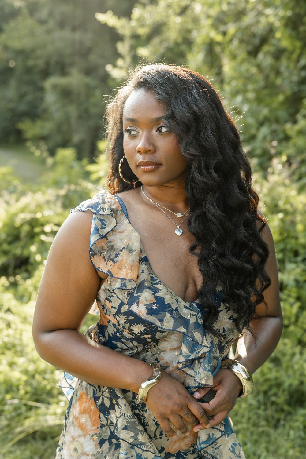 A person in a floral print dress poses outdoors against a green natural background on a sunny day.