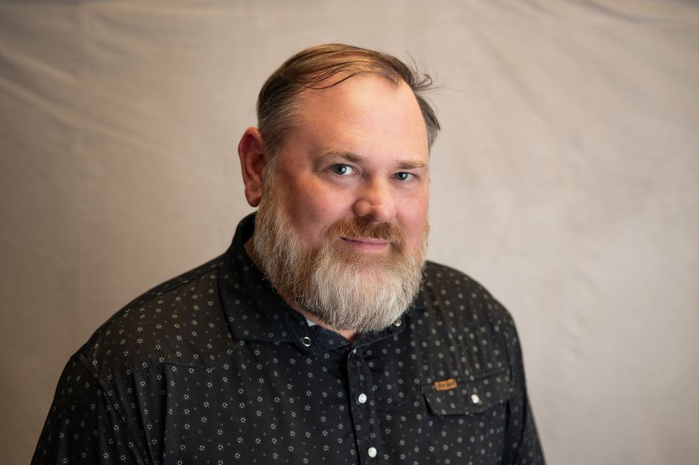 Person with full beard and dark polka dot shirt smiles while sitting against light background.