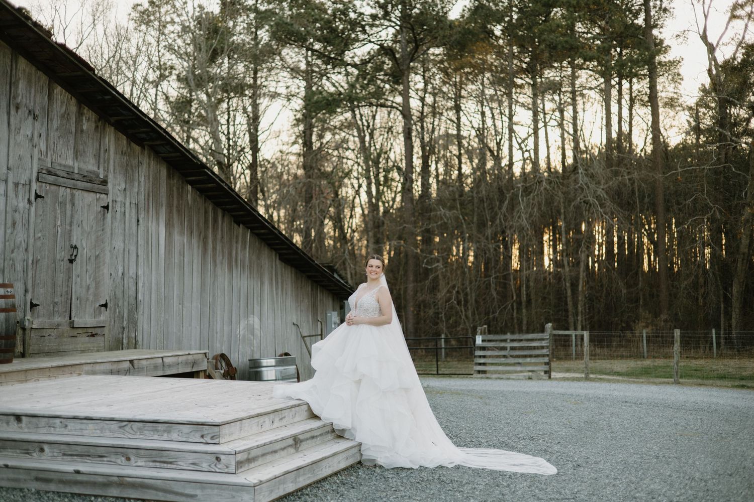 A flowing white wedding dress trails on rustic wooden steps beside a weathered barn at sunset in a pine forest.