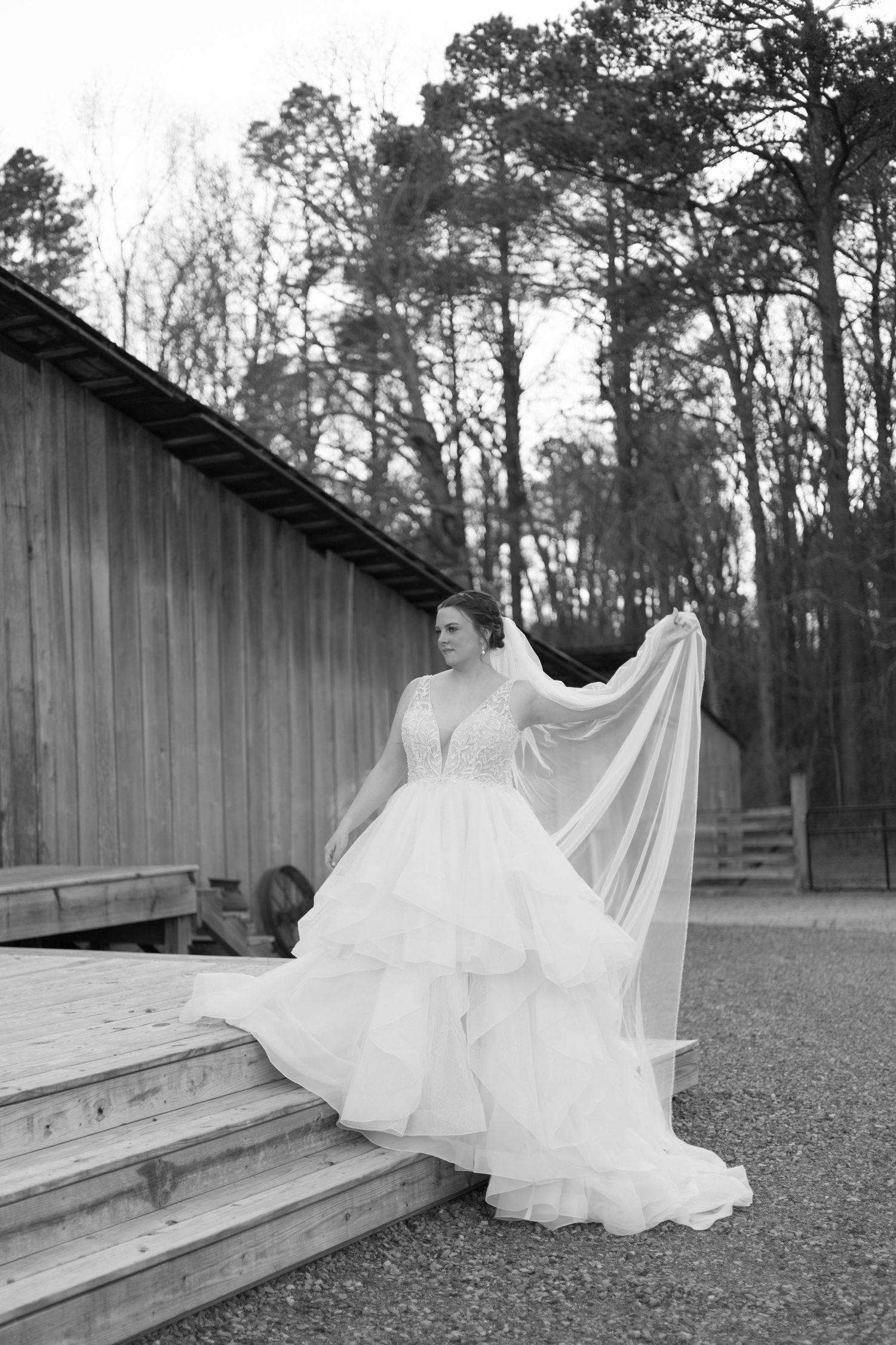 A bride in a flowing white ballgown poses gracefully on wooden steps outside a rustic barn surrounded by tall trees.