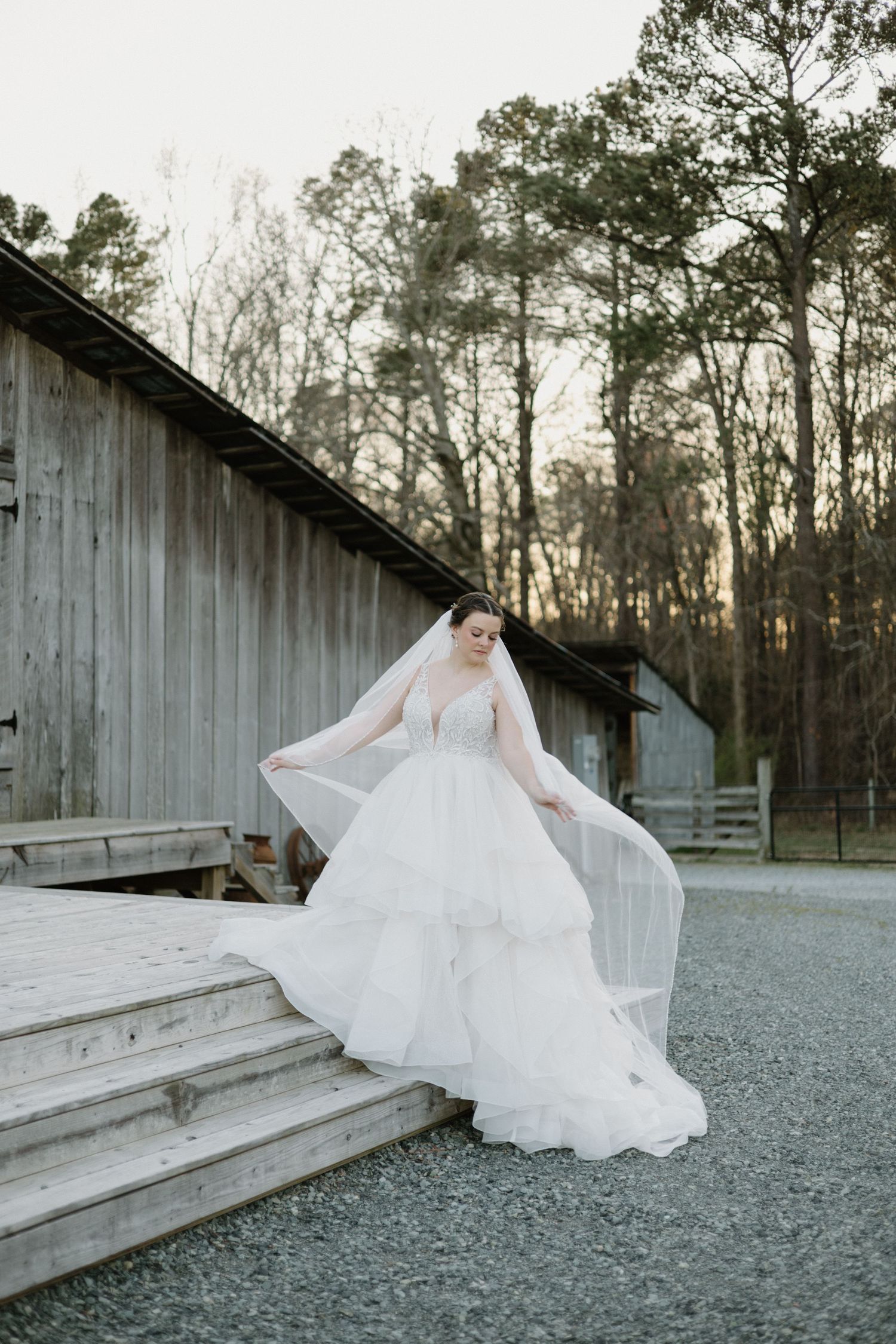 A person in a flowing white wedding dress poses on wooden steps against a rustic barn and forest backdrop at dusk.